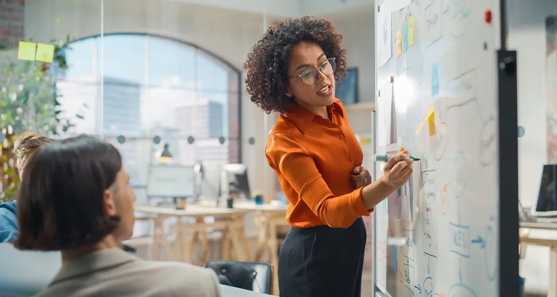 A woman in an orange shirt writes on a whiteboard covered with notes and diagrams, presenting to colleagues in a modern, sunlit office; two people are seated, listening attentively, with large windows and city buildings visible in the background.