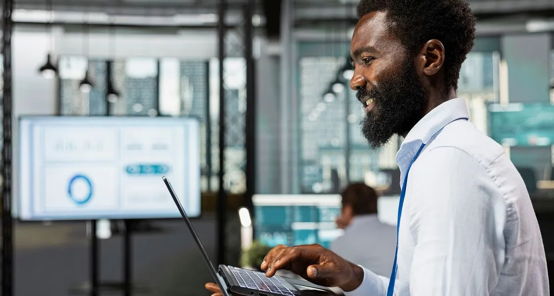 A man with a beard, wearing a blue lanyard and blue shirt, smiles while using a laptop in a modern office. In the background, blurred figures work at desks and a screen displays charts and graphs. The setting appears professional and high-tech.