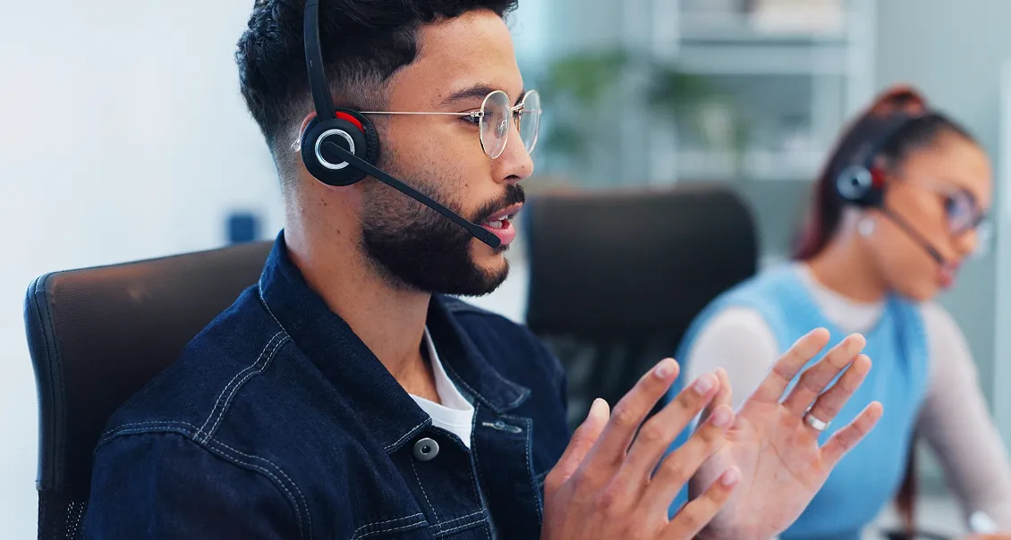 A man with a beard and glasses wearing a headset and denim jacket is speaking and gesturing with his hands at a desk. In the background, a woman with a ponytail and headset is also seated, working at a computer. Both appear to be in a modern office.
