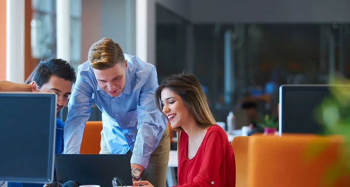 Three young adults, two men and one woman, smile and collaborate around a laptop in a modern office with bright natural light. Computer monitors and orange chairs are visible, creating a vibrant, professional atmosphere.