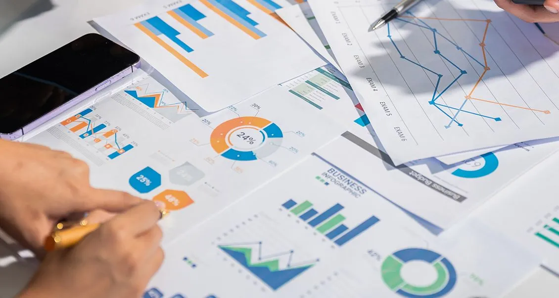 A close-up of hands reviewing colorful business charts and graphs on paper at a desk; visible are bar graphs, pie charts, and line graphs, along with a smartphone and a pen, suggesting data analysis or financial planning.