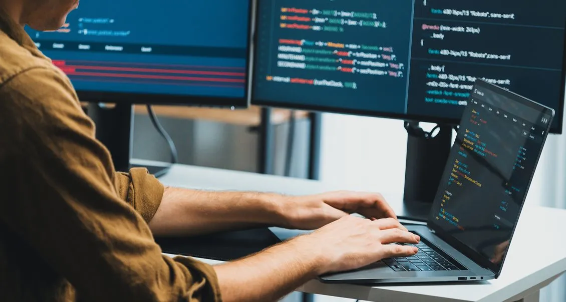 A person types on a laptop at a desk, with two monitors displaying code in various colors. The individual wears a brown shirt and works in a modern, well-lit office environment, focusing on programming tasks.