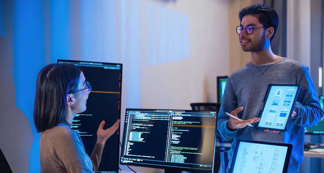Two people in an office discuss software displayed on computer monitors. One person sits facing code on screens, gesturing, while the other stands, holding a tablet with app wireframes visible. The room is lit with blue and neutral tones, suggesting a tech workspace.