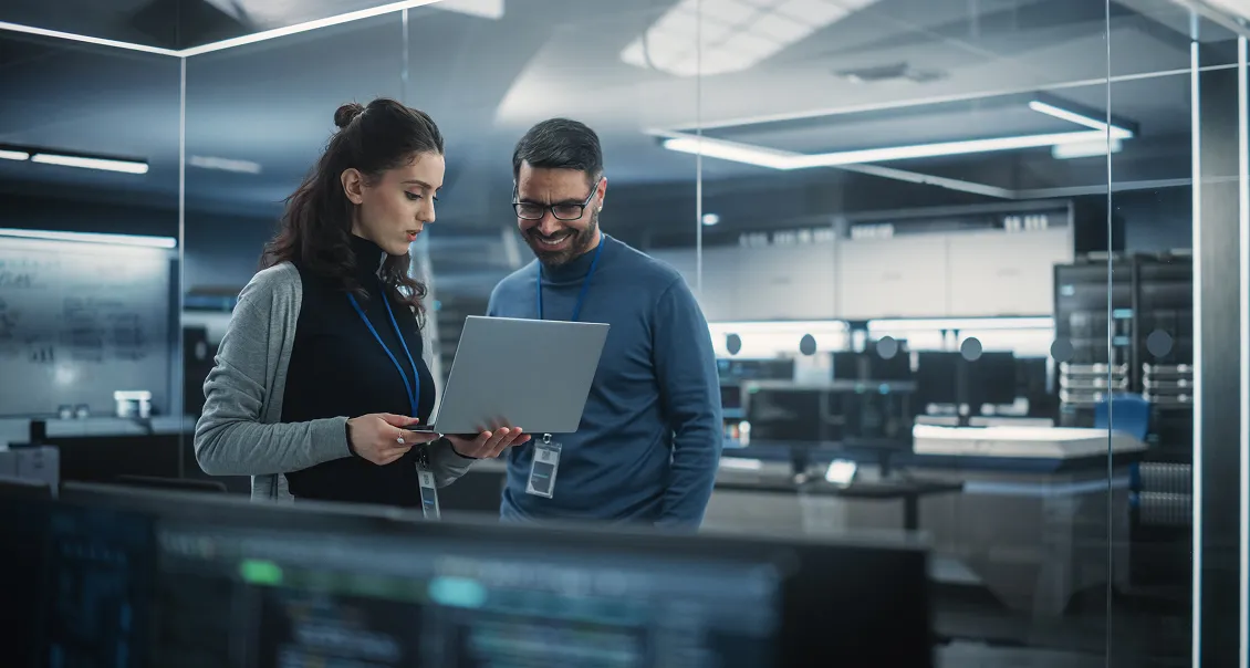 Two professionals stand in a modern, glass-walled office with computer monitors and tech equipment. One woman holds a laptop, showing it to a smiling man. Both wear ID badges and business-casual clothes, collaborating in a high-tech environment.