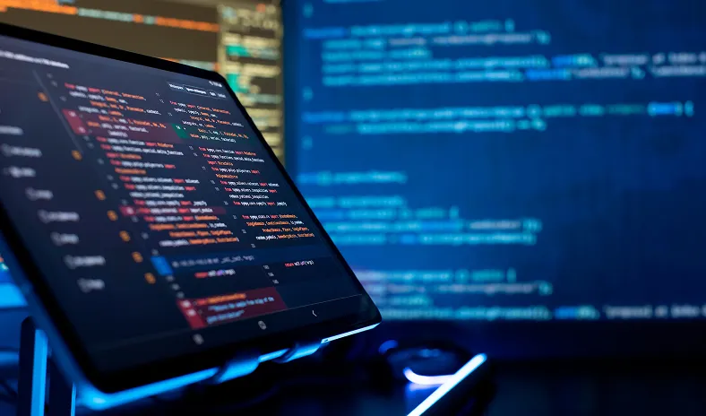 A tablet on a stand displays lines of colorful code in a dark room, with a large monitor in the background also showing blue programming code. The lighting is dim, highlighting the technology and programming environment.