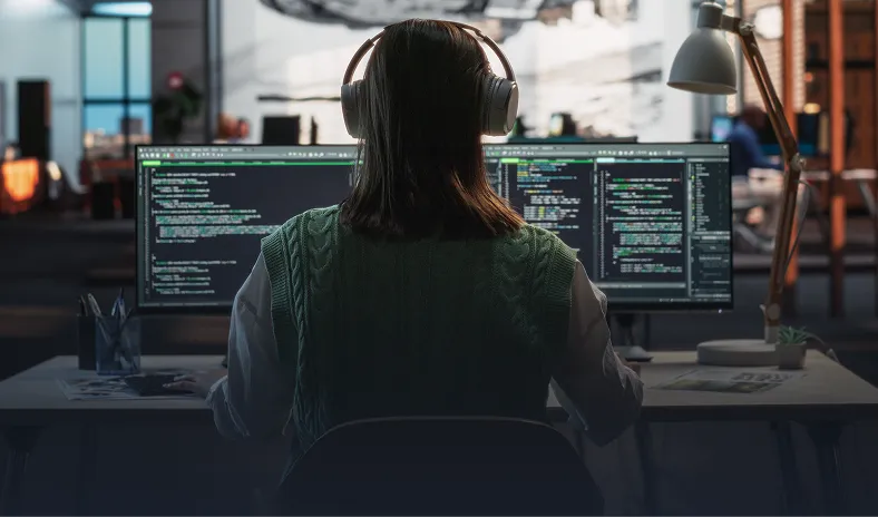 A person with long hair, wearing headphones and a green sweater, sits at a desk in a dimly lit office, working on two large computer monitors displaying code. The workspace includes a lamp, a plant, and office supplies, with a blurred office in the background.