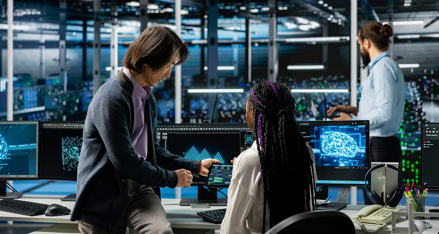 Three people work in a modern, high-tech office with many monitors showing data and digital graphics. Two are talking by a desk covered in screens, while a third stands in the background, facing large server racks and computer equipment.