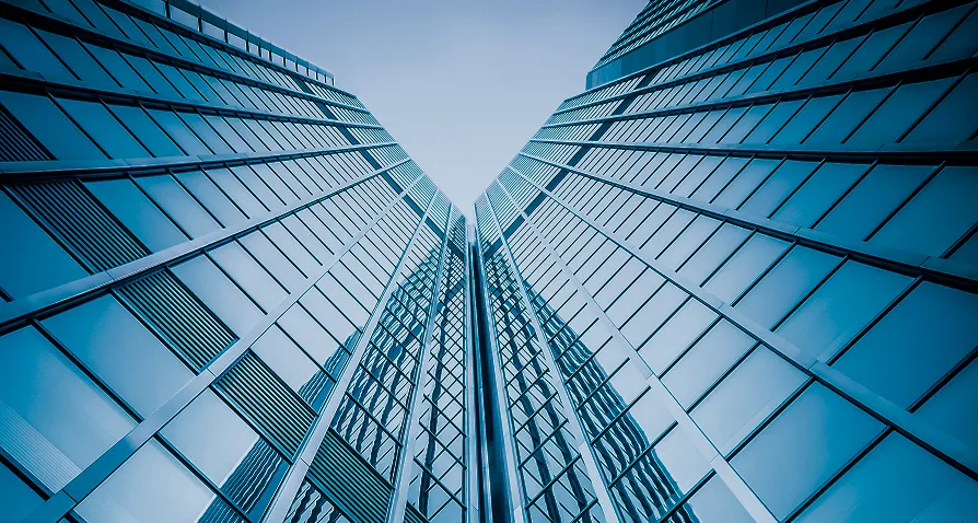 Upward view of two tall modern glass skyscrapers converging at a sharp angle, with reflective blue windows and a clear sky above, creating a symmetrical, geometric perspective.