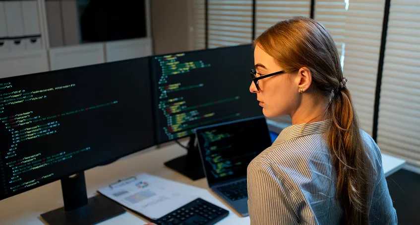 A woman with long brown hair and glasses, wearing a striped shirt, sits at a desk facing dual monitors displaying lines of code. She looks over her shoulder. A laptop, keyboard, and documents are on the desk. Blinds cover the window behind her.