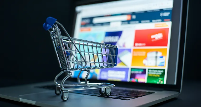 A small toy shopping cart sits on a laptop keyboard in front of a screen displaying a colorful online shopping website with various product categories and promotions, symbolizing online shopping or e-commerce.