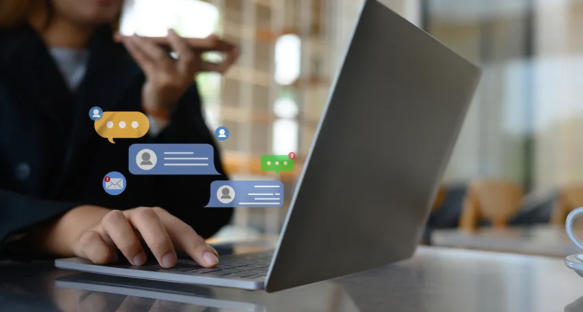 A person using a laptop at a table, with one hand typing and the other holding a phone near their mouth. On-screen digital chat and email notification icons are overlaid. The background is softly blurred, suggesting a modern indoor setting.