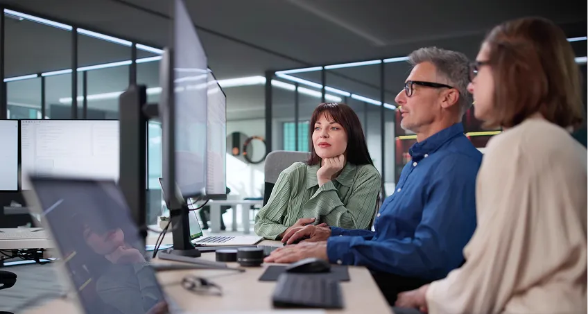 Three people sit closely together at an office desk, focused on large computer monitors. The man in the center types on a keyboard, while two women on either side look attentively at the screens. The modern office has glass walls and ample daylight.