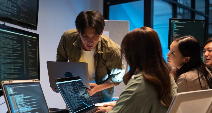 Four young adults in a modern office work on laptops displaying code. One person stands, leaning over with a laptop, discussing something with the others, who are seated and listening attentively. Large monitors with code fill the background.