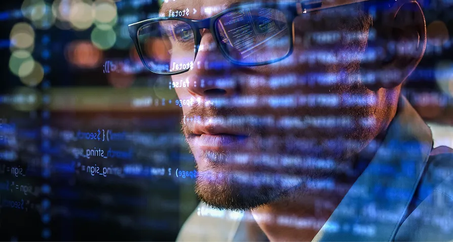 A man wearing glasses intently looks at a computer screen filled with lines of blue and white code, which are reflected in his glasses. The background is blurred with colorful lights, highlighting his focus on programming.