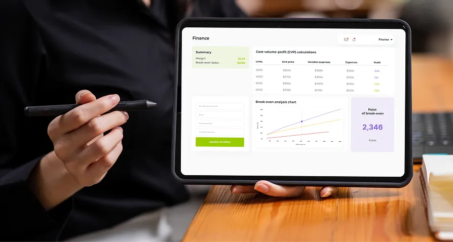A person holding a tablet with a stylus. The tablet screen displays a finance dashboard with charts, tables, and a highlighted Point of Break-even: 2,346. Most of the display is taken up by a break-even analysis graph and related data.