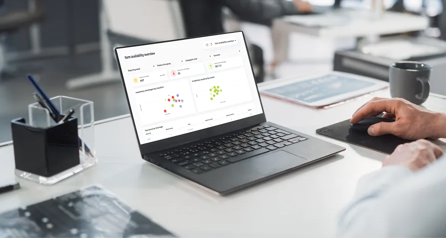 A person uses a wireless mouse next to a laptop displaying a data analytics dashboard with graphs and charts. The laptop sits on a white desk with office supplies, a pen holder, and blurred figures in the background, suggesting a modern office environment.