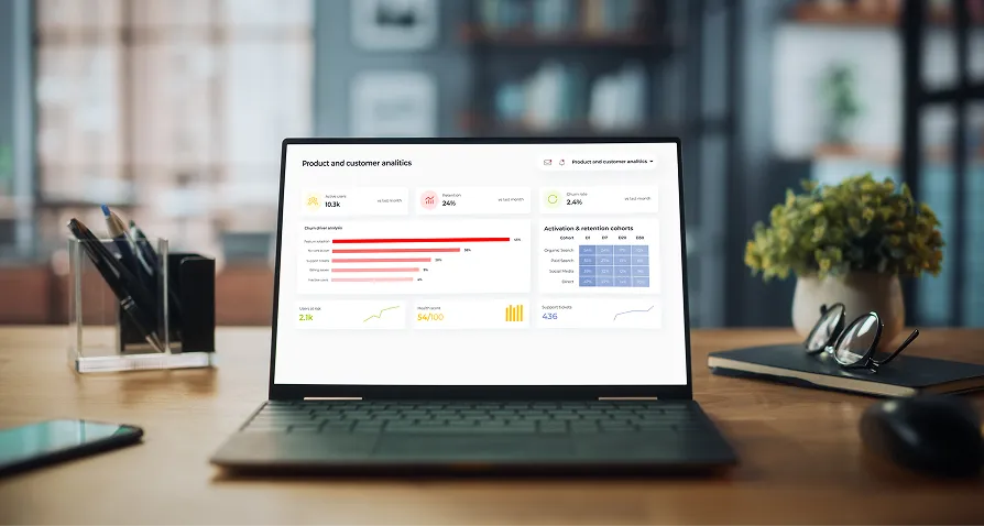A laptop on a wooden desk displays a product and customer analytics dashboard with charts, graphs, and data. Items on the desk include a plant, pen holder, glasses, and notepad, with shelves, books, and decor visible in the blurred background.