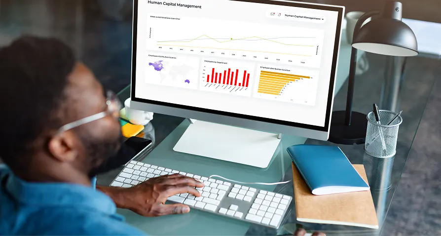 A man sits at a desk using a desktop computer, viewing charts and graphs related to human capital management on the screen. The desk holds a keyboard, mouse, pen, closed blue notebook, eyeglasses, and a lamp. The setting appears to be an office.