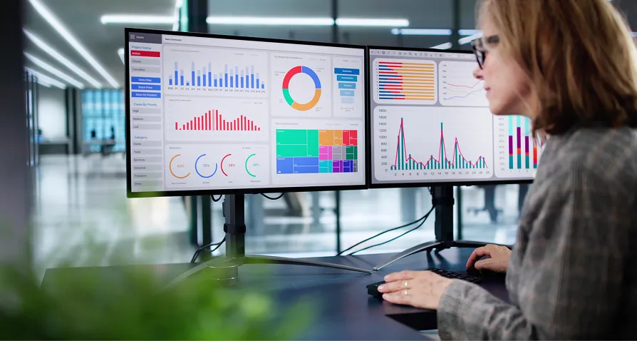 A woman wearing glasses sits at a desk, analyzing colorful graphs and charts displayed on two computer monitors. The screens show bar charts, pie charts, and line graphs. The office space in the background is modern and brightly lit.