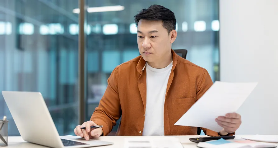 A man sits at a desk holding papers in one hand and using a laptop with the other. He wears a brown jacket over a white shirt and appears focused, working in a modern office with glass walls in the background.