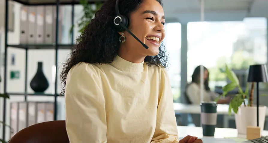 A woman with curly hair wearing a headset and a light yellow blouse smiles while sitting at a desk in a modern office. A coffee cup, green plant, and another person working are visible in the background. Sunlight brightens the space.