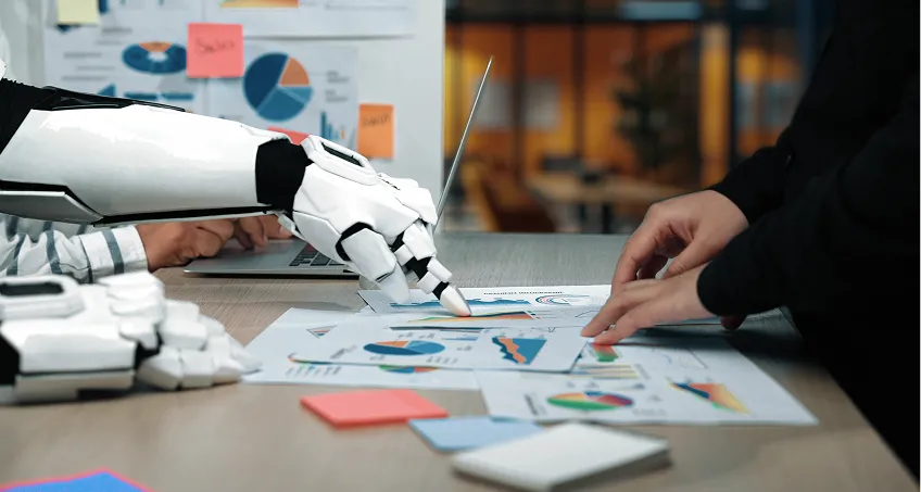 A robot hand and a human hand point to charts and graphs on a desk covered with papers, laptops, and sticky notes in an office setting, symbolizing collaboration between artificial intelligence and humans in data analysis and business.