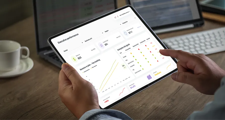 A person holds a tablet displaying business analytics dashboards with charts, graphs, and financial data. The tablet is on a wooden desk near a laptop, a keyboard, and a coffee cup, suggesting a work or office environment.