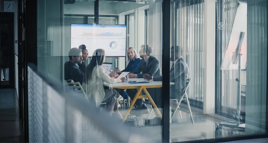 Seven people in business attire sit around a conference table in a glass-walled meeting room, engaged in discussion. A large screen behind them shows charts and graphs. The scene is modern and professional, with daylight streaming through blinds.