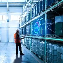 A worker in a hard hat and orange safety vest stands in a warehouse aisle, holding a laptop. Large blue barrels are stacked on metal shelves. A transparent digital display shows analytics and charts, indicating inventory or stock data.