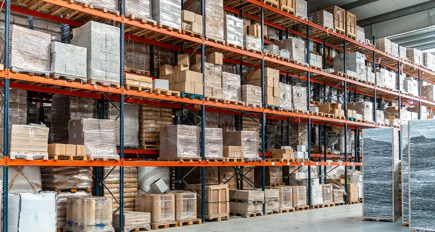 A large warehouse with tall metal shelves stacked with various boxes and packages, some wrapped in plastic. The shelves are organized into multiple rows and levels, filled with goods stored on wooden pallets. The floor is clean and spacious.