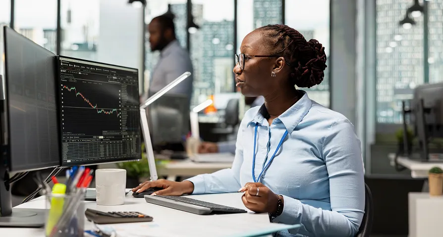 A woman wearing glasses and business attire works at a desk with two monitors displaying stock charts in a modern office with large windows. She uses a mouse and keyboard, with office supplies, coffee mug, and blurred colleagues in the background.