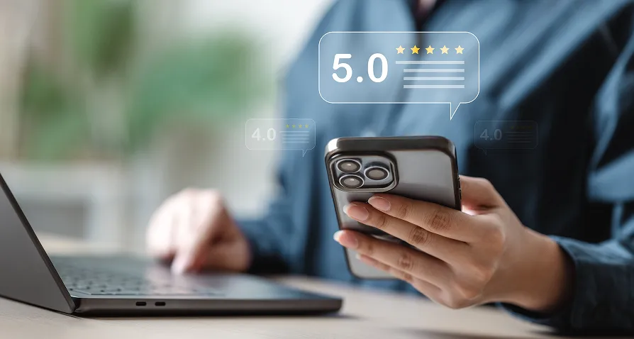 A person holds a smartphone in one hand and uses a laptop with the other. Floating icons show a 5.0-star rating above the phone and 4.0 ratings in the background, suggesting online reviews or feedback. The person’s face is not visible.