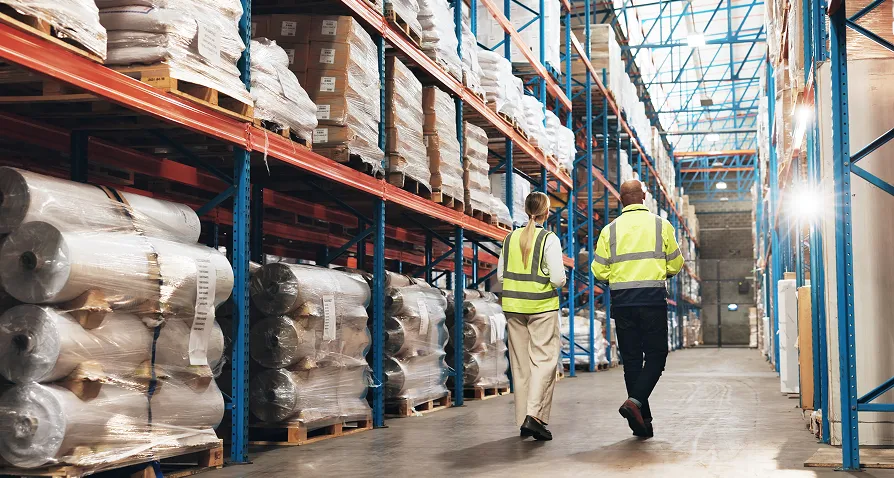 Two workers wearing yellow safety vests walk side by side down an aisle in a large warehouse. Tall racks on both sides store goods wrapped in plastic. The warehouse is brightly lit, spacious, and organized for industrial storage.