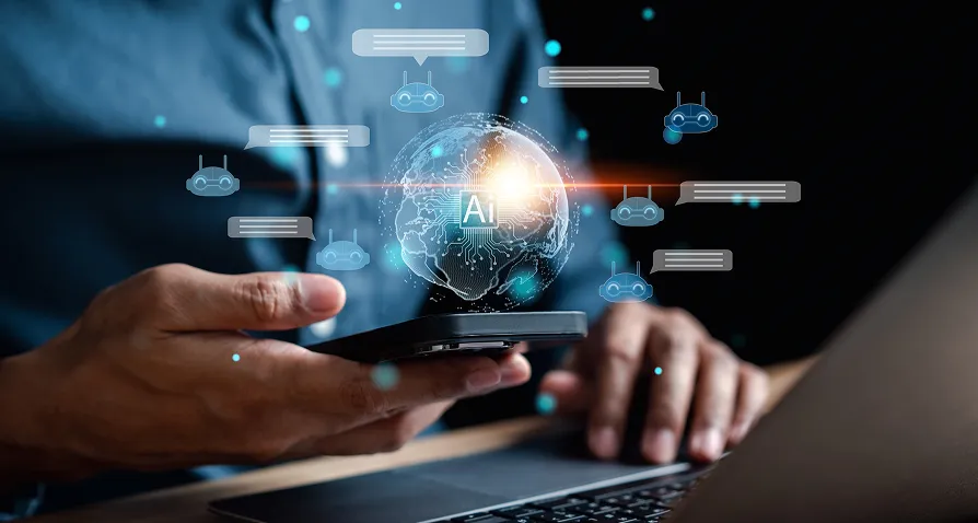 A person holds a smartphone in one hand with a laptop nearby. Above the phone, a glowing digital globe labeled AI is surrounded by chat bubbles and robot icons, symbolizing artificial intelligence and virtual communication.