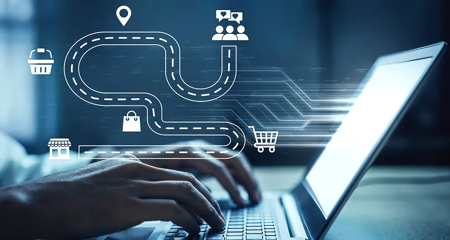 Hands typing on a laptop keyboard with a digital illustration overlay showing a winding path connecting icons of a store, shopping bag, shopping cart, location pin, and group of people, symbolizing a digital customer journey.