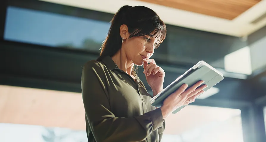 A woman with dark hair tied back stands indoors, holding a tablet. She looks thoughtful, with one hand touching her lips and the other holding the device. She wears a green blouse and is surrounded by sunlight streaming through large windows.
