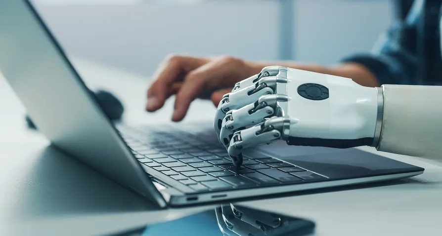 A close-up of a robotic hand and a human hand typing together on a laptop keyboard. The scene suggests collaboration between artificial intelligence and humans, with a smartphone lying on the desk beside the laptop.