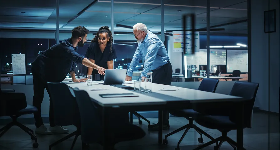 Three colleagues stand around a conference table in a modern office at night, discussing something on a laptop. The room is dimly lit, with large windows and office supplies on tables in the background. All appear engaged and collaborative.