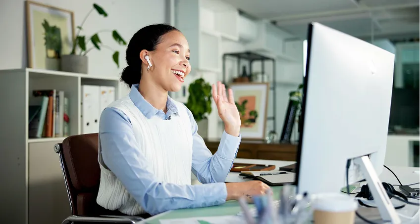 A woman sits at a desk in a bright office, smiling and waving at a large computer monitor. She wears wireless earbuds, a light blue shirt, and a white sweater vest. Plants, shelves, and folders are in the background, suggesting a modern workspace.