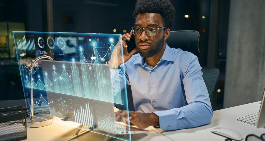 A man wearing glasses and a blue shirt sits at a desk in a modern office at night, talking on a smartphone. In front of him is a transparent digital display showing financial charts, graphs, and data visualizations.