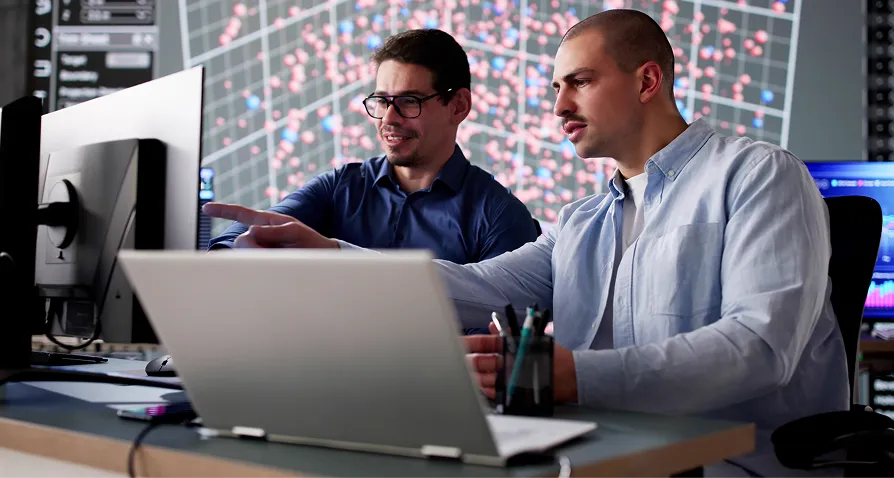 Two men sit at a desk with a laptop and desktop monitor, discussing something on the screen. Both appear focused; one points at the monitor. In the background, a large digital display shows colorful data points on a grid, suggesting a tech or research setting.