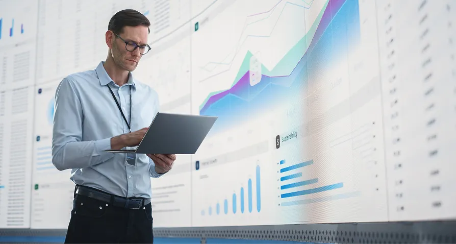A man wearing glasses and business attire stands using a laptop. Behind him is a large wall screen displaying various colorful charts, graphs, and data visualizations, indicating analysis or monitoring of information.
