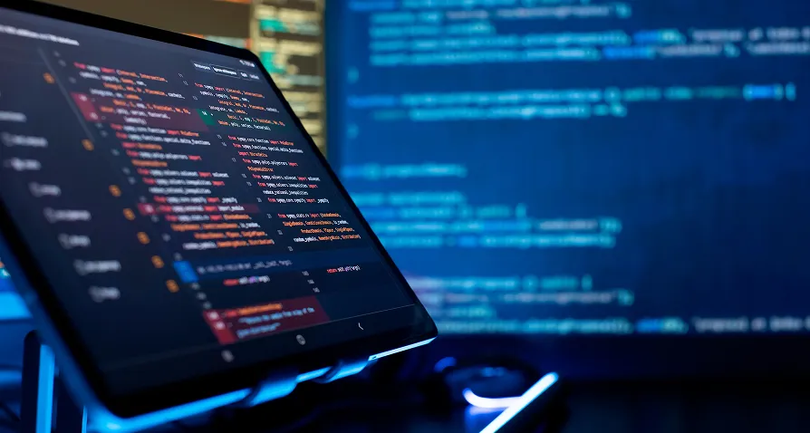 A tablet displays colorful lines of code in a dark room. In the background, a large screen shows blurred blue and white programming code. The lighting is low, emphasizing the tech-focused workspace.