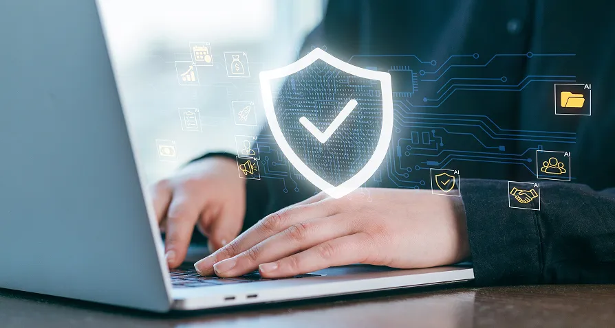 A person types on a laptop. Overlaid graphics show a glowing shield with a checkmark, symbolizing cybersecurity or data protection, with icons of locks, documents, and gears floating nearby, suggesting secure digital activity.