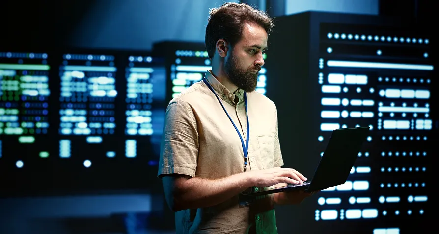A man with a beard, wearing a beige shirt and a lanyard, stands in a server room. He is typing on a laptop. Behind him are rows of illuminated server racks displaying blue and green lights, indicating an advanced tech environment.