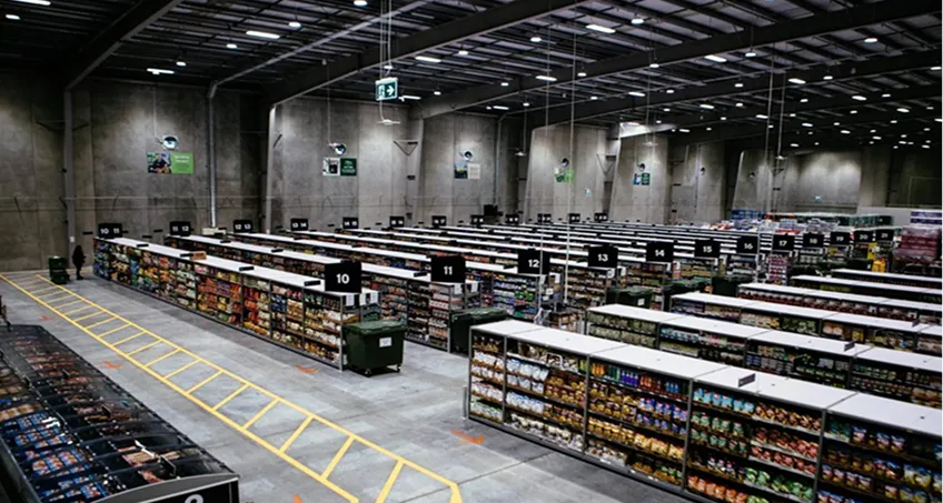 A large warehouse-style grocery store with high ceilings, concrete walls, and bright lighting. Multiple aisles are filled with various packaged goods, each labeled with black signs numbered 10 to 15. Yellow lines mark pathways on the polished floor.